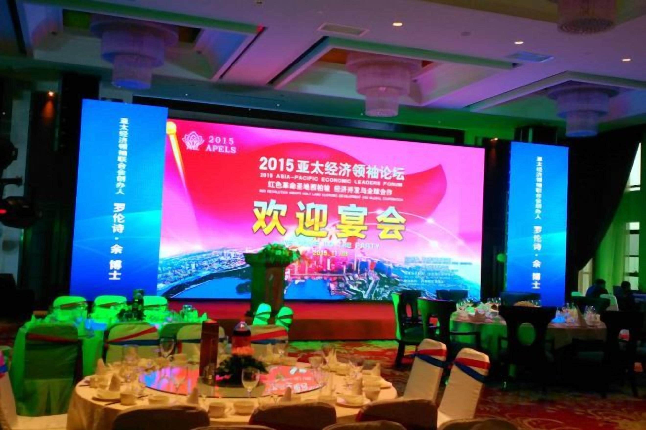 A conference room with a large screen displaying event information, round tables with chairs, and a stage with a floral arrangement.