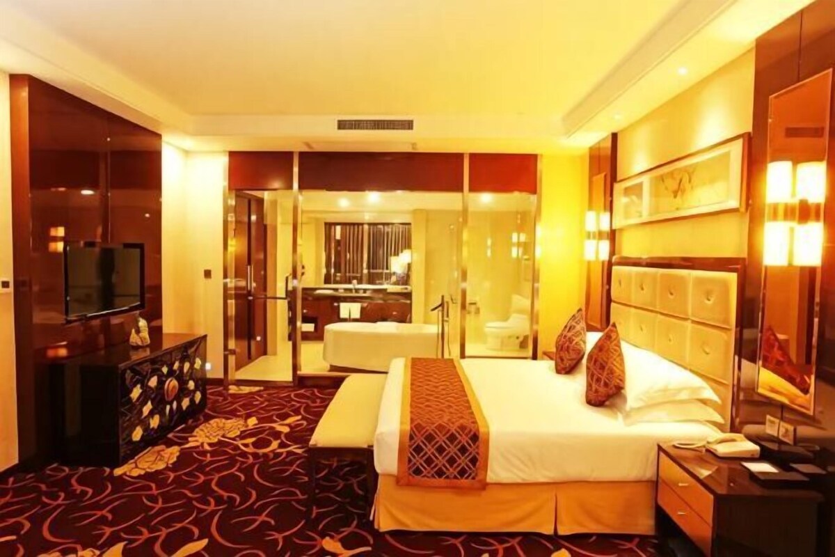 Habitación de hotel con cama, mesita de noche, televisión y baño visible a través de una puerta de vidrio.
