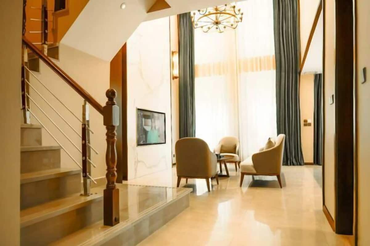 Een trap met houten spijlen die leidt naar een plateau, een kroonluchter en twee fauteuils in een goed verlichte kamer.