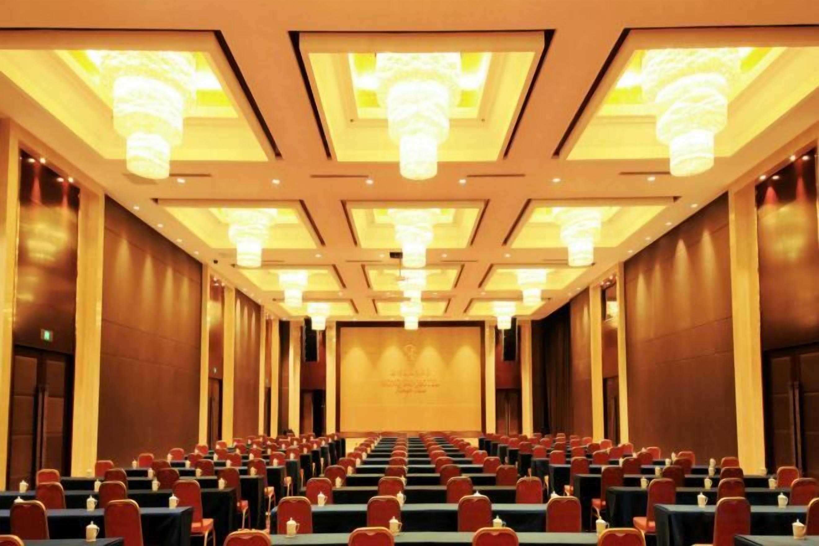 Une salle de conférence avec des rangées de tables et de chaises, une scène à l’avant et des lustres au plafond.