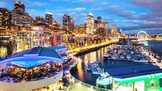 Un paisaje urbano frente al agua, con un puerto deportivo, edificios altos y una estructura circular distintiva con techo azul.