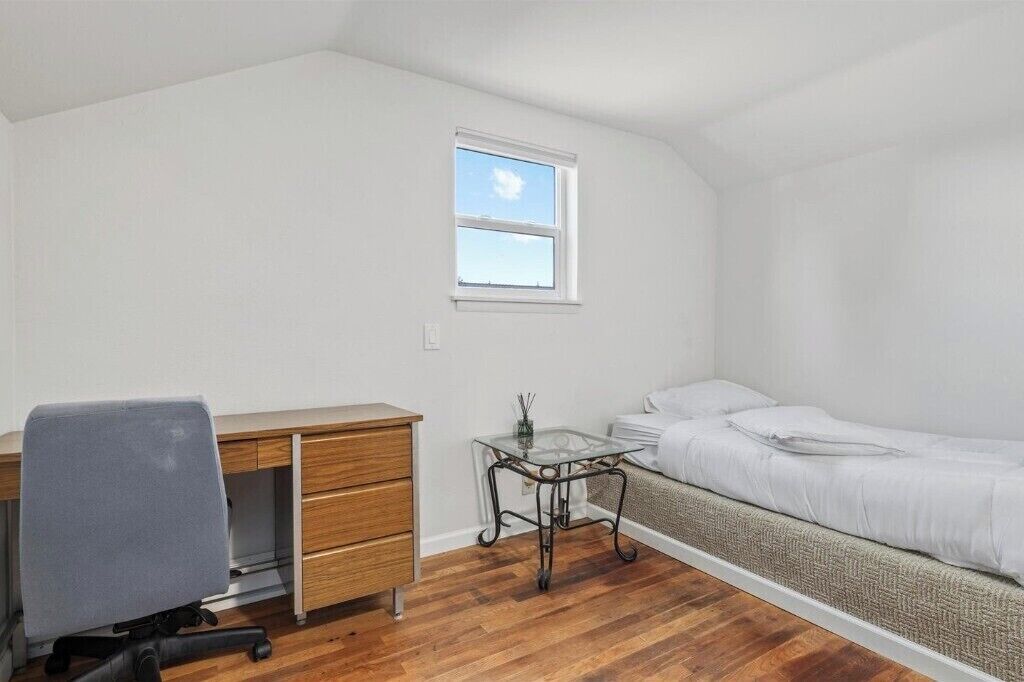A small, well-lit room with a wooden desk, a chair, a bed with a white mattress, and a window with a view of the sky.