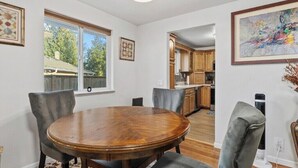 Un comedor con una mesa redonda de madera y cuatro sillas grises tapizadas. Se aprecia una ventana con vistas a un patio trasero y árboles. Las paredes son blancas y hay un aplique en el techo.
