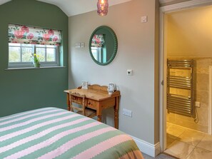 A bedroom with a bed, a wooden nightstand, a mirror, a window with floral curtains, and a bathroom with a towel rack. - Escape to this picturesque retreat in Salton, near Malton. (Salton, near Malton)