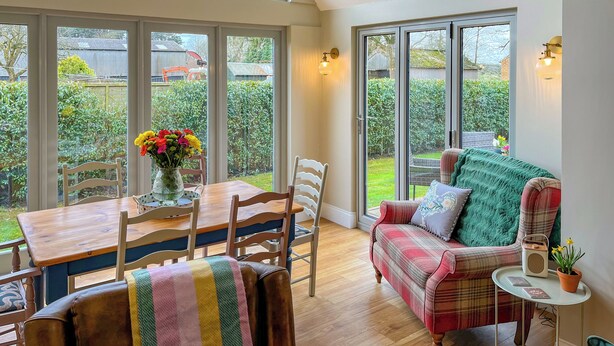 A bright room with a wooden dining table, chairs, and a sofa with a plaid blanket. There are large windows and a view of a garden. - Escape to this picturesque retreat in Salton, near Malton. (Salton, near Malton)
