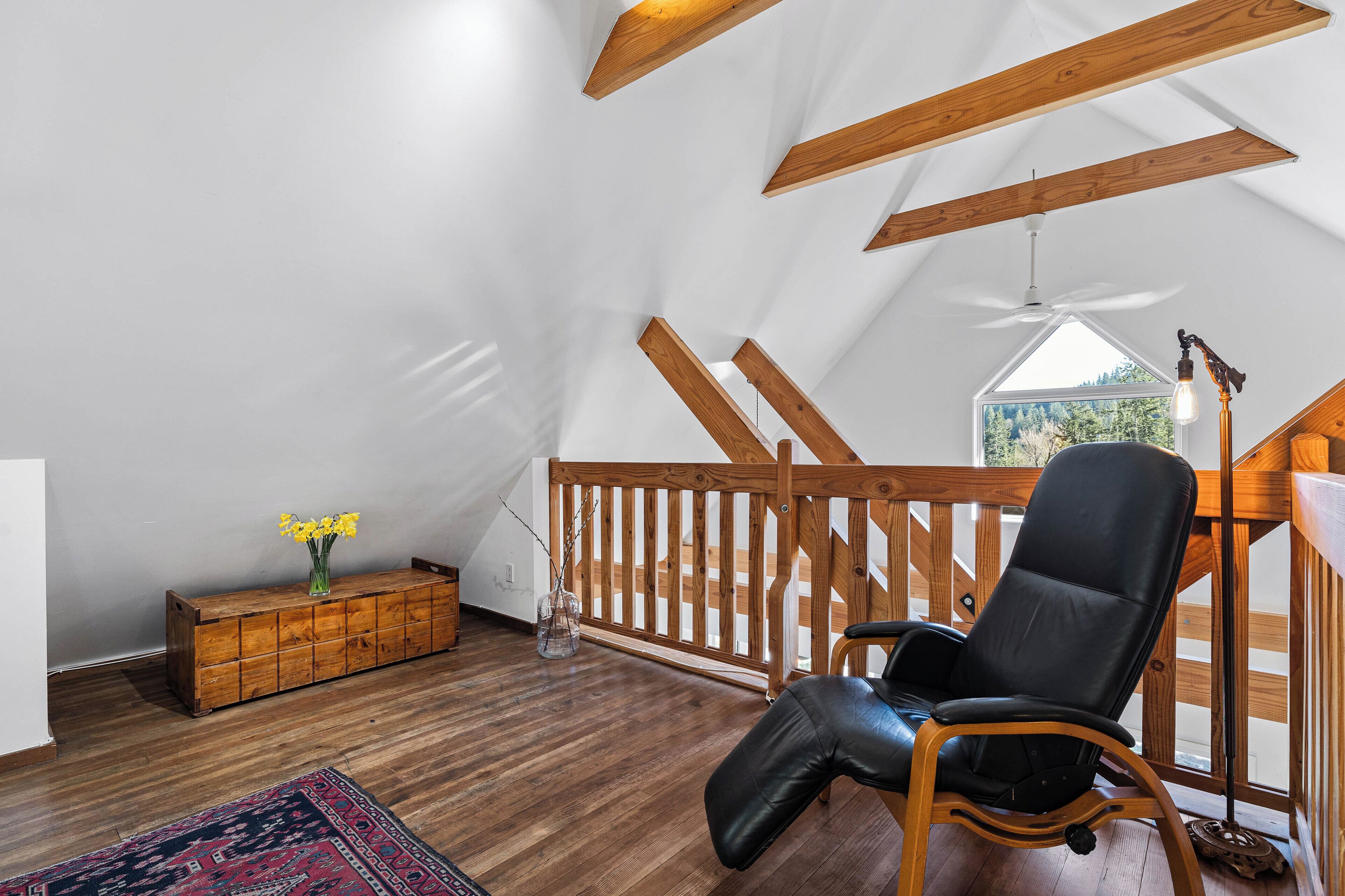 A loft bedroom with wooden beams, a black leather chair, a wooden dresser, and a view of the outdoors.