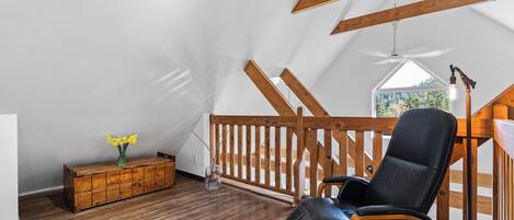 A loft bedroom with wooden beams, a black leather chair, a wooden dresser, and a view of the outdoors.