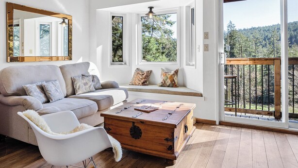 Un salon spacieux avec un plafond haut, des poutres en bois, un fauteuil à bascule, un canapé et une vue sur une forêt, visible à travers de grandes fenêtres.