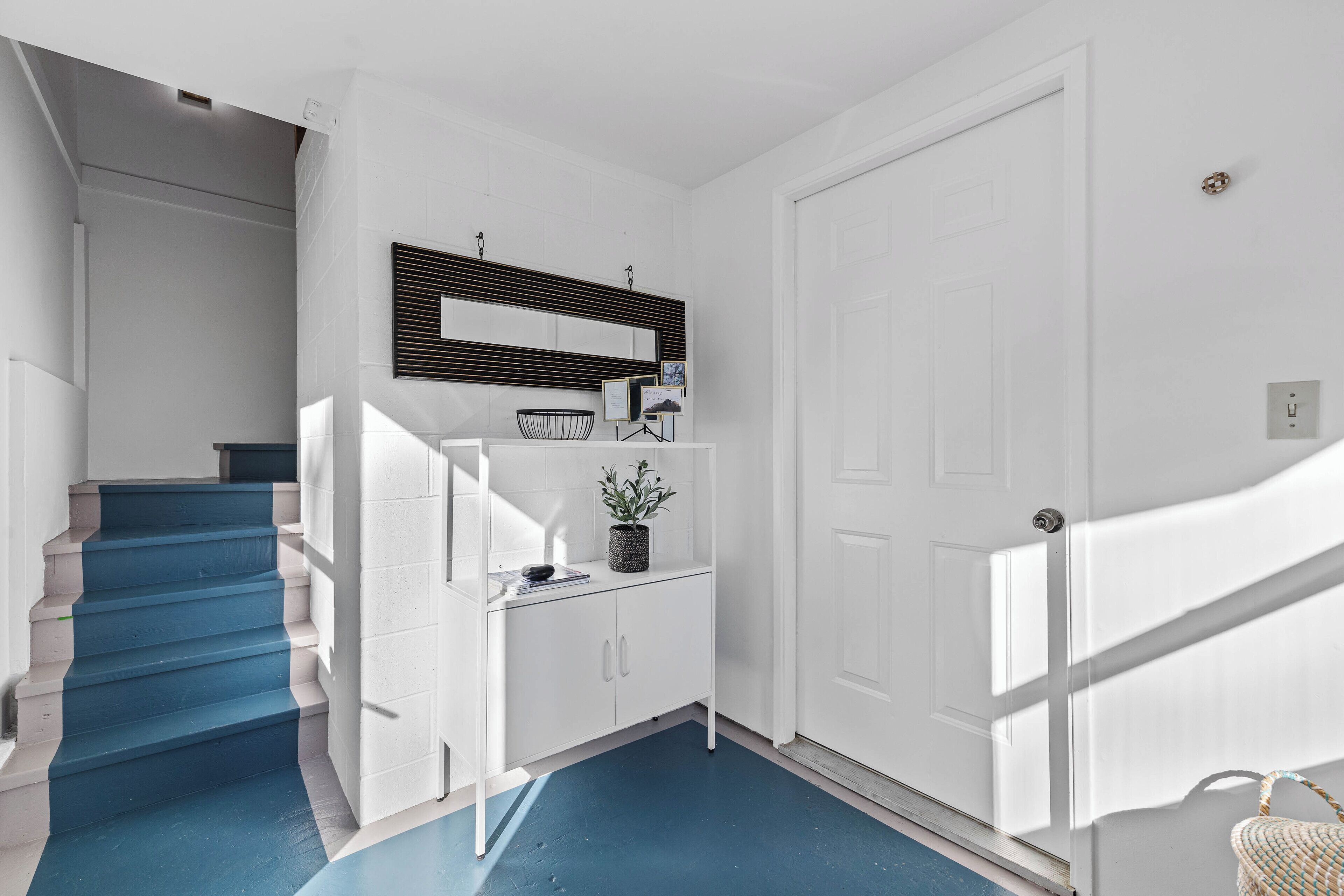 A staircase with blue steps leading to a landing with a white wall, a black-framed mirror, and a small potted plant.