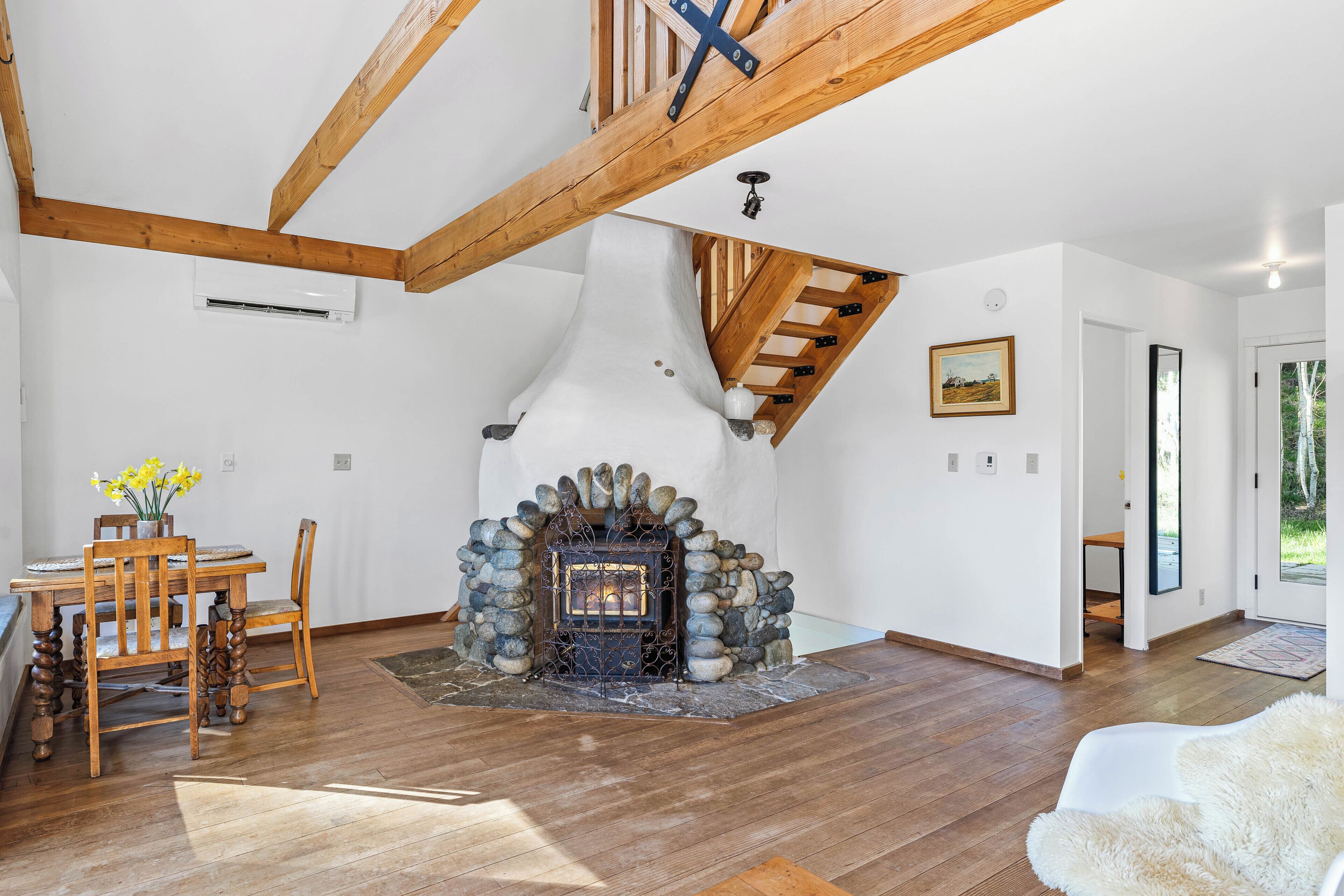 A spacious living room with a stone fireplace, wooden beams, a dining area with a table and chairs, and a view of the outdoors.
