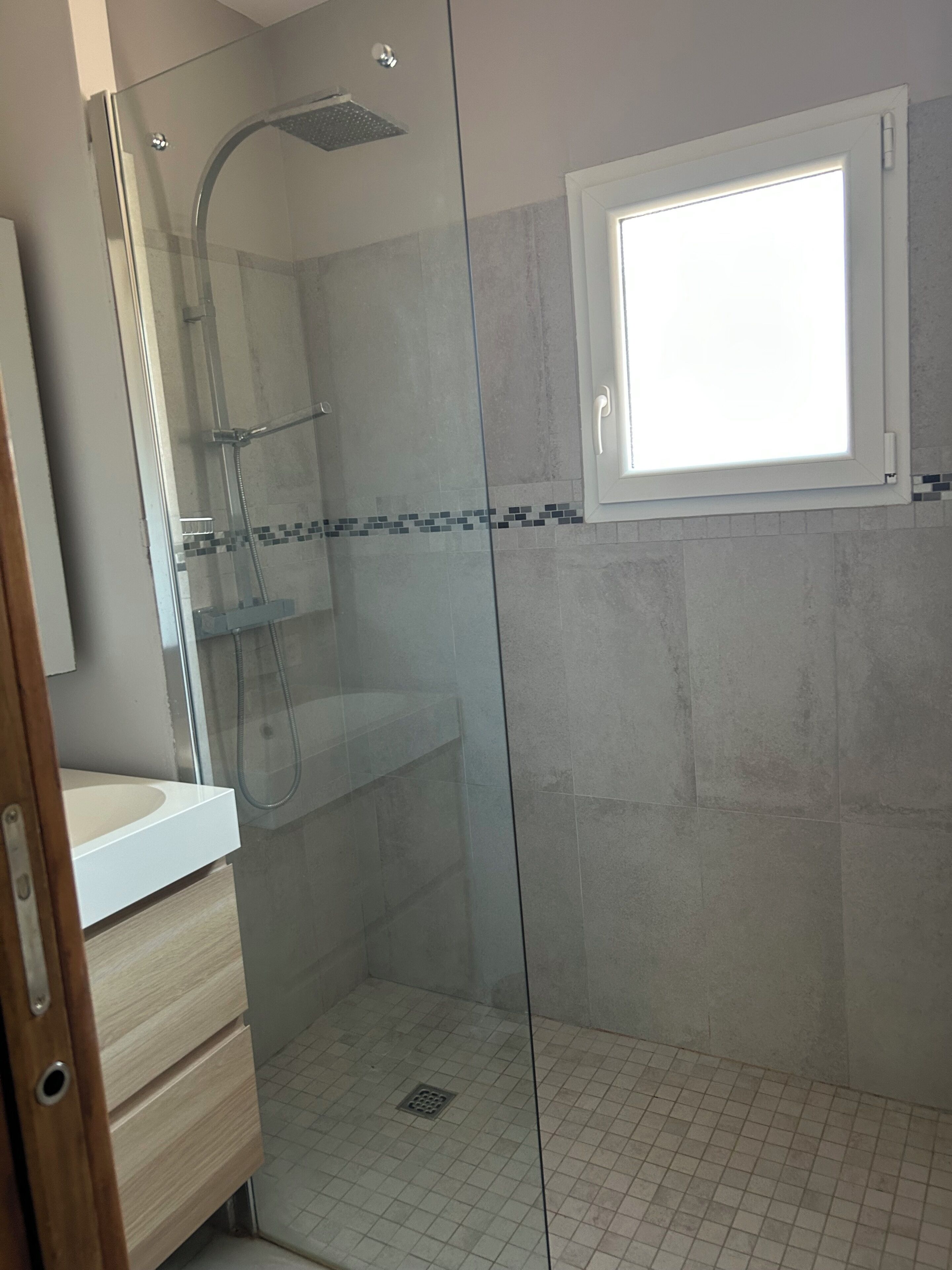 Une salle de bain avec une douche à parois de verre, un lavabo blanc et une fenêtre située au-dessus de la douche.