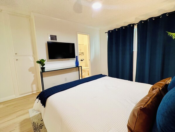 Un dormitorio con una cama, un televisor sobre un mueble, una planta y una ventana con cortinas.