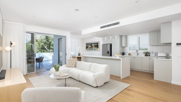 Una cocina moderna con una isla central, un comedor con sofá blanco y mesa, y vistas al exterior a través de un ventanal corredero.