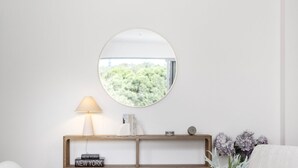 Un espejo redondo en una pared, una lámpara de noche, una consola de madera con libros y un reloj, y un florero con flores.