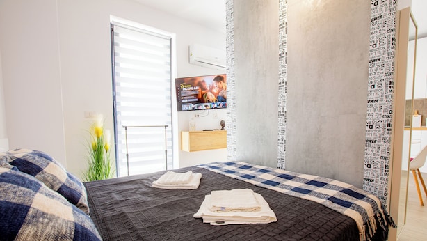 A bedroom with a bed, a TV, and a window with blinds. - ATENEO - Studio DeLuxe MODERN Apartment, BIG Balcony, Plus FREE Parking (Timișoara)