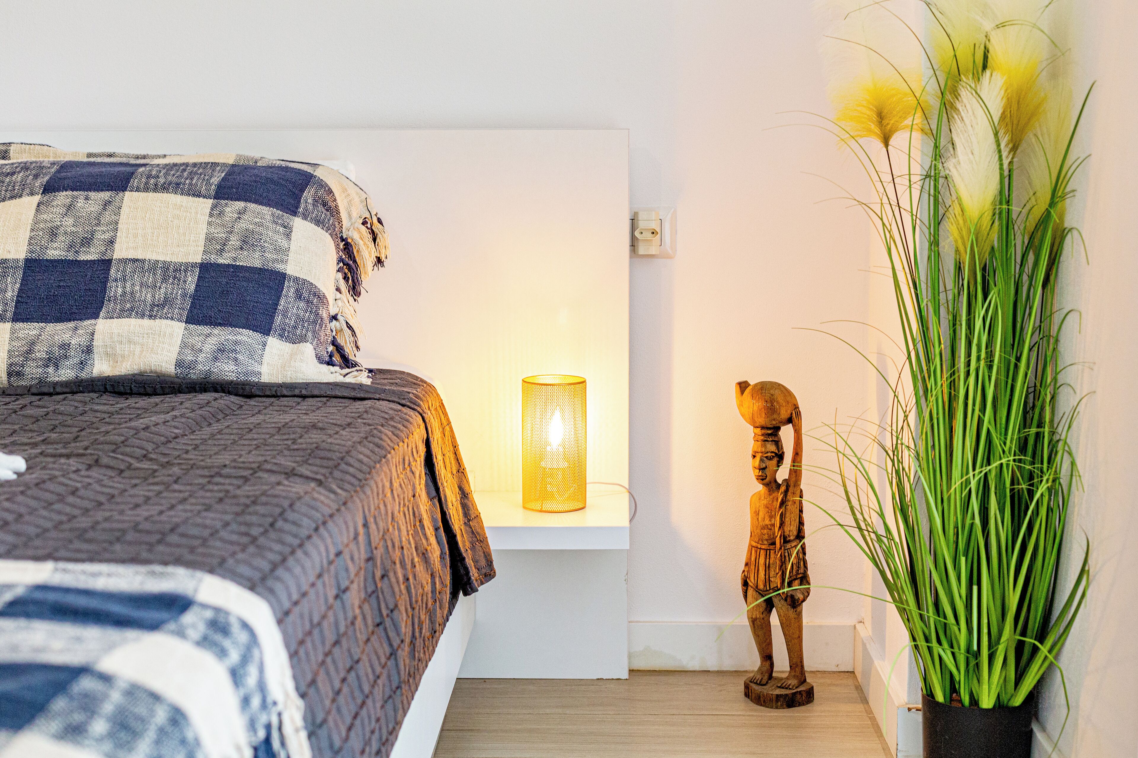 Un dormitorio con colcha a cuadros, una lámpara de noche, una escultura de madera y una planta en maceta.