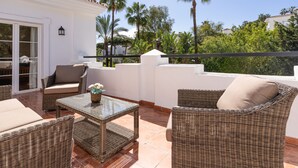 A balcony with wicker furniture, a glass table, and a view of palm trees and buildings.