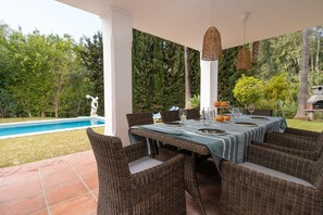 An outdoor dining area with wicker chairs and a table set for six, overlooking a swimming pool and lush greenery.