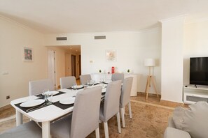 Un coin repas avec une table blanche dressée pour quatre personnes, des chaises grises, une petite table avec une lampe et un téléviseur posé sur un meuble.