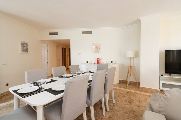 Un comedor con una mesa blanca para cuatro personas, sillas grises, una mesita con una lámpara y un televisor sobre un mueble.