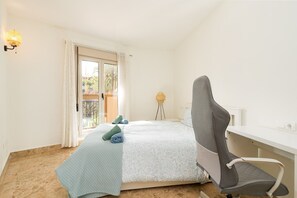 Un dormitorio con cama, escritorio y silla. Hay una ventana con cortinas, una lámpara y vistas al exterior.