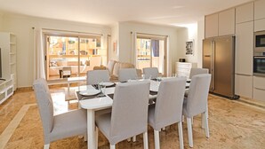 Eine moderne Küche mit Einbauofen und Kühlschrank, ein Essbereich mit weißem Tisch und grauen Stühlen sowie ein Balkon mit Blick auf Gebäude.