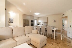 Una sala de estar moderna con un sofá seccional color beige, un comedor con mesa y sillas blancas, y una cocina con gabinetes y electrodomésticos blancos.