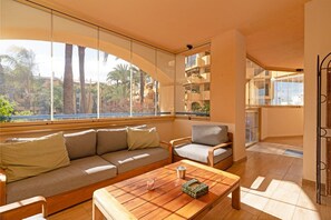 Una sala de estar moderna con un sofá, sillas, una mesa de centro y ventanas grandes que ofrecen vista a palmeras y edificios.