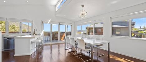 A modern kitchen with a dishwasher, a dining area with a white table and chairs, and large windows offering views of the outdoors.