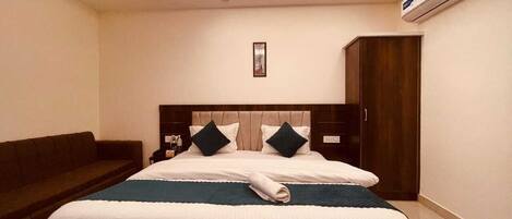 A hotel room with a bed, a dark-colored sofa, a wooden wardrobe, and an air conditioning unit.