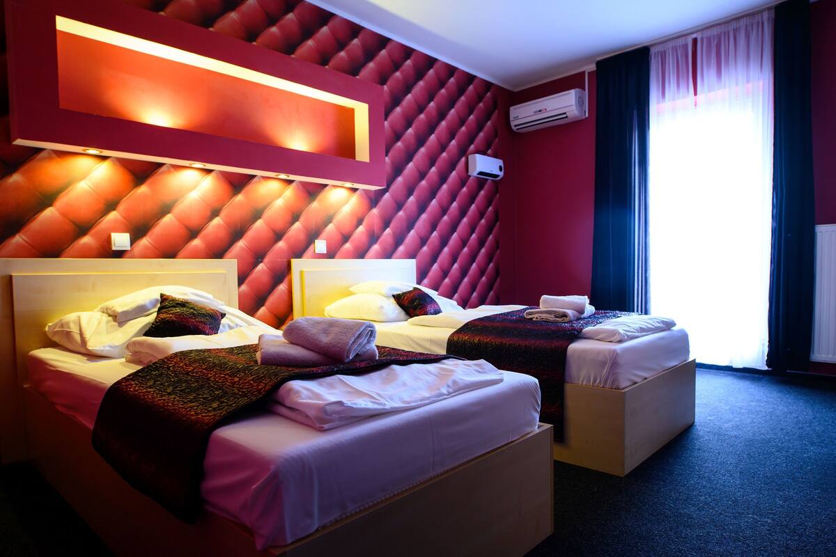 A hotel room with two beds, a red wall with a diamond pattern, and a window with curtains.