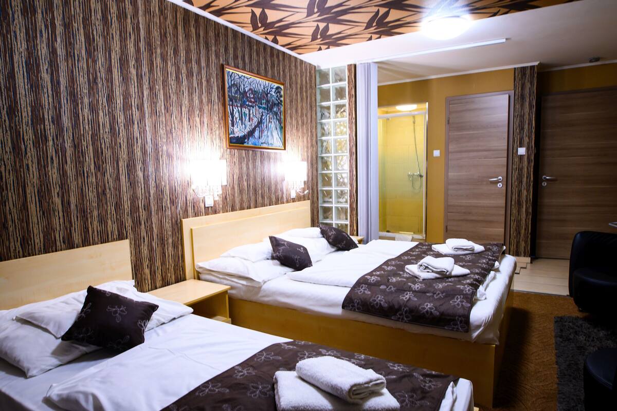 A hotel room with two beds, a wooden wall, a painting, and a bathroom visible through a glass door.