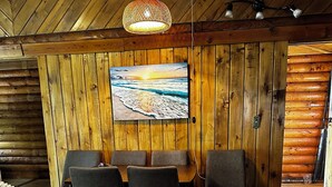 Un interior de madera con una mesa de comedor y sillas, una lámpara colgante y un cuadro que representa una escena de playa.