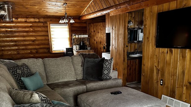 Un salón acogedor con un sofá modular, un televisor de pantalla plana, paredes de madera y una cocina al fondo.
