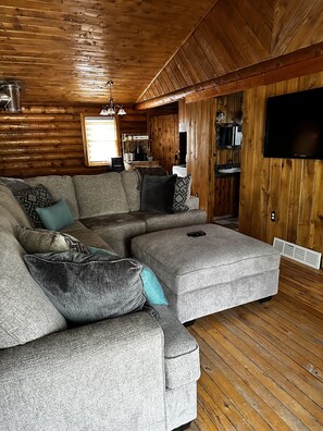 Een gezellige woonkamer met een hoekbank, een flatscreen televisie, houten wanden en een keuken in de achtergrond.