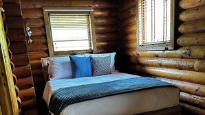 Una habitación de cabaña de madera con una cama, dos ventanas y piso de madera.