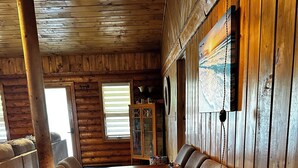 Interior de una cabaña de madera con una amplia mesa de comedor rodeada de sillas, techo de madera y una ventana con persianas.