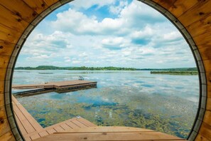 Una estructura de fusta circular amb un pont de fusta que s'estén sobre l'aigua, envoltada de vegetació i un cel nuvolós.