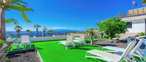 A rooftop pool area with artificial green turf, white lounge chairs, and a view of the sea and mountains.