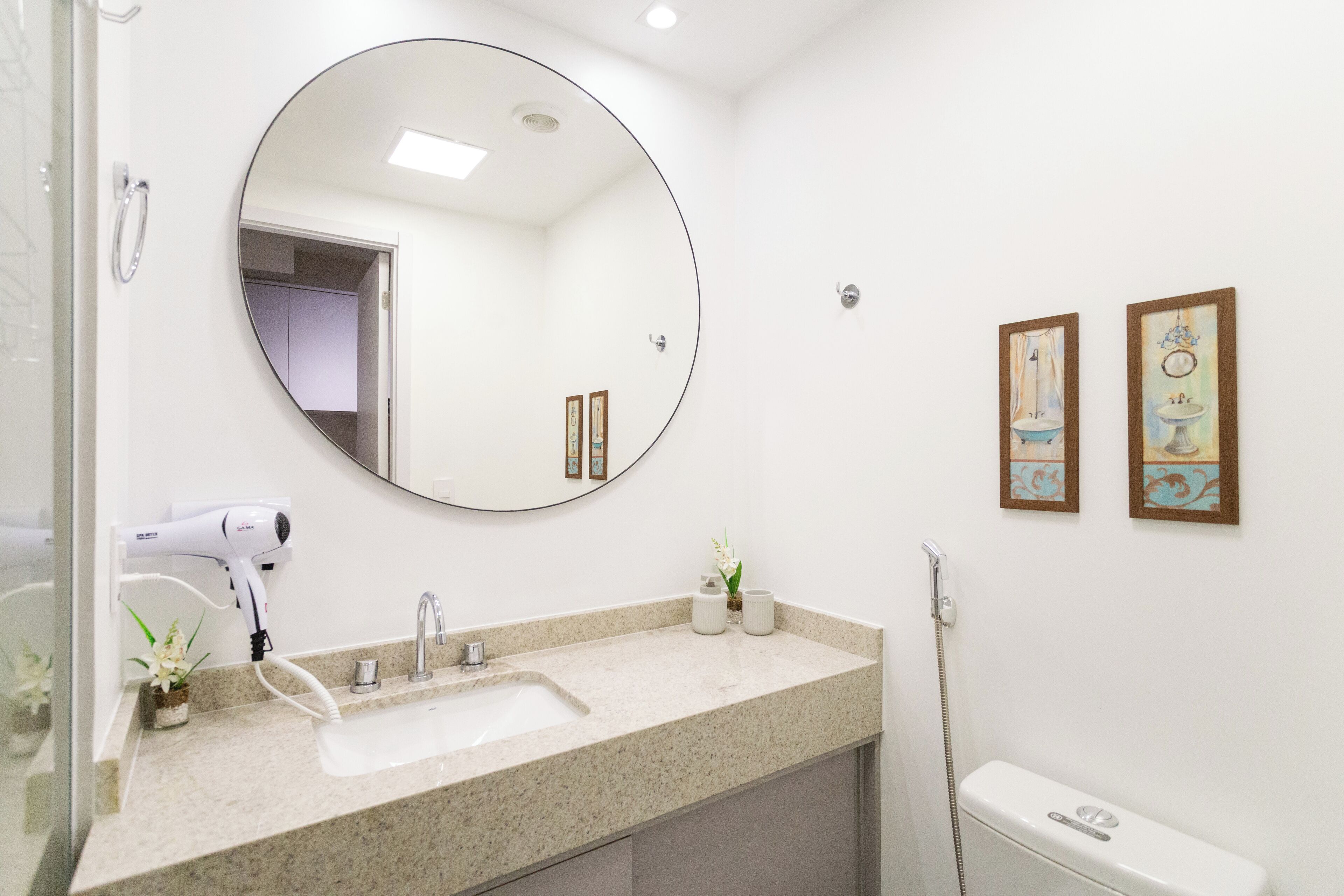 Un baño con un espejo redondo, un lavabo, un secador de pelo, un inodoro y dos cuadros en la pared.