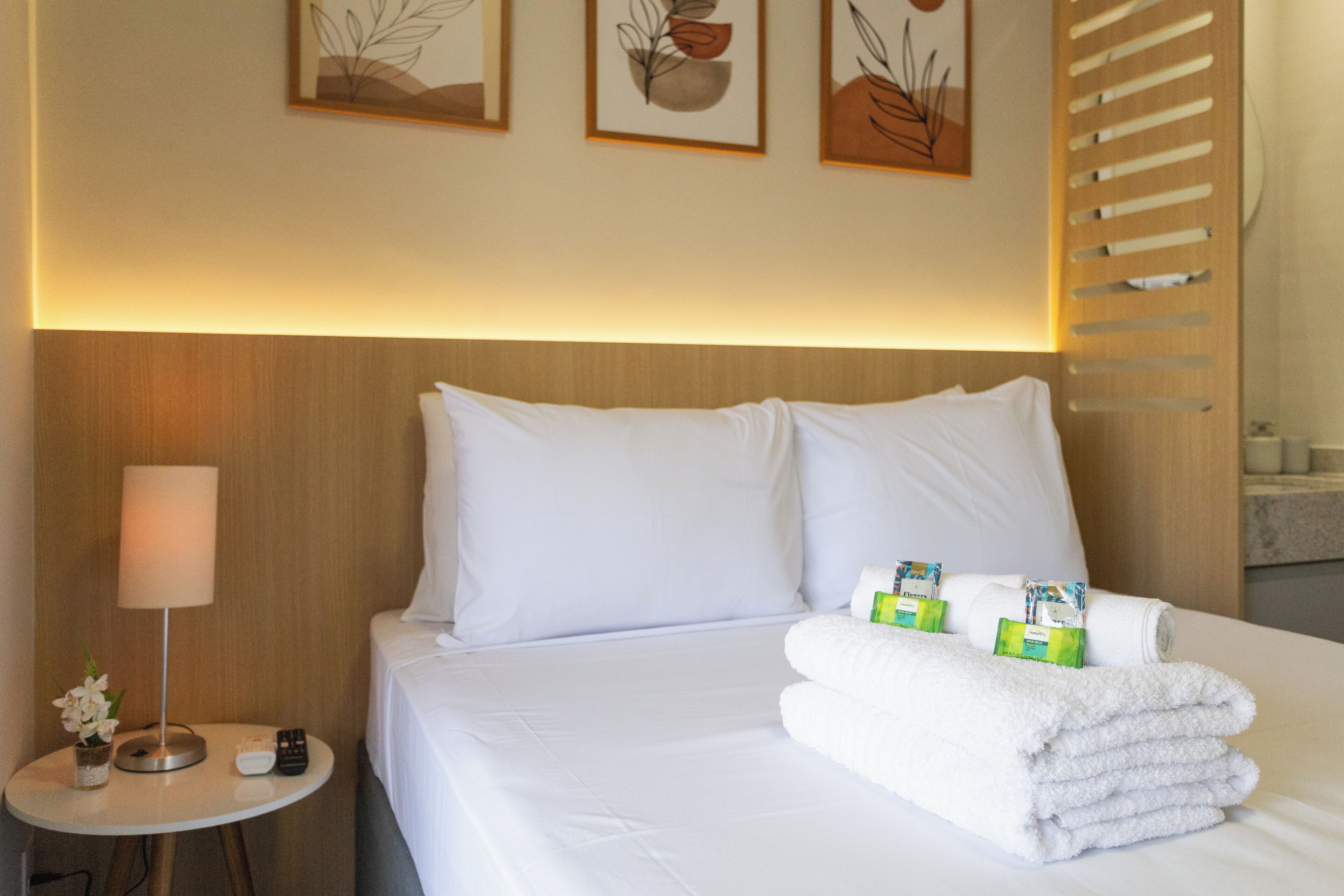 A neatly made bed with white linens, a small bedside table with a lamp, and a towel rack with towels.
