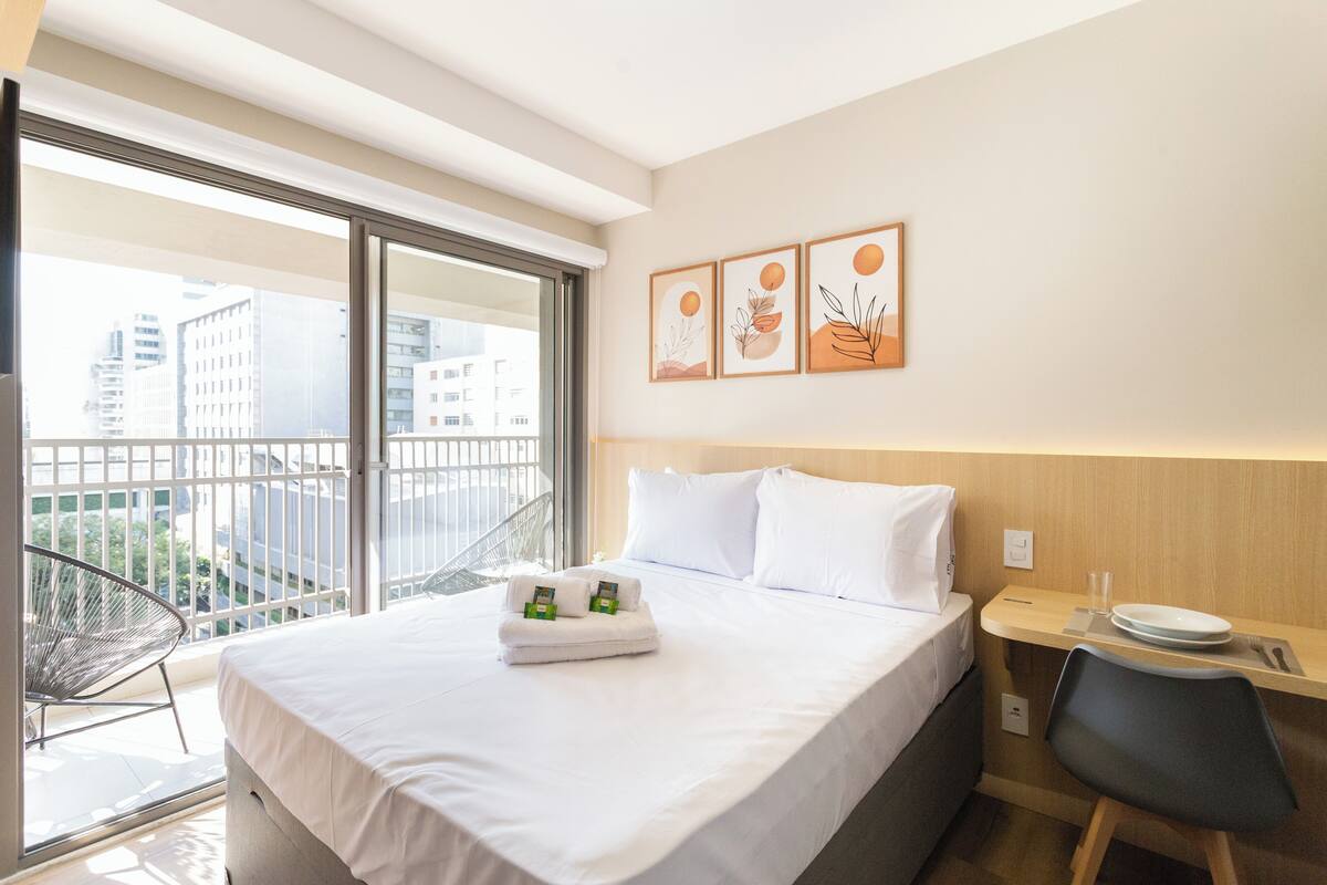 A hotel room with a bed, a desk, a chair, and a balcony with a view of buildings.