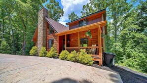 A modern log cabin with a stone chimney, a wooden deck, and a surrounding forest.