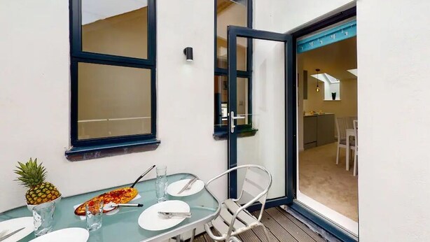 Een glazen tafel met borden, bestek en een ananas op een balkon met een witte muur en een raam.