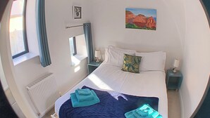 Een slaapkamer met een bed, raam, radiator, schilderij en nachtkastje.