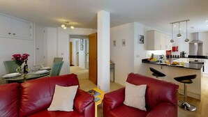 Un appartement moderne doté d’un salon avec des canapés en cuir rouge, d’un coin repas avec une table dressée pour quatre personnes et d’une cuisine équipée d’appareils électroménagers en acier inoxydable et d’un plan de travail noir.
