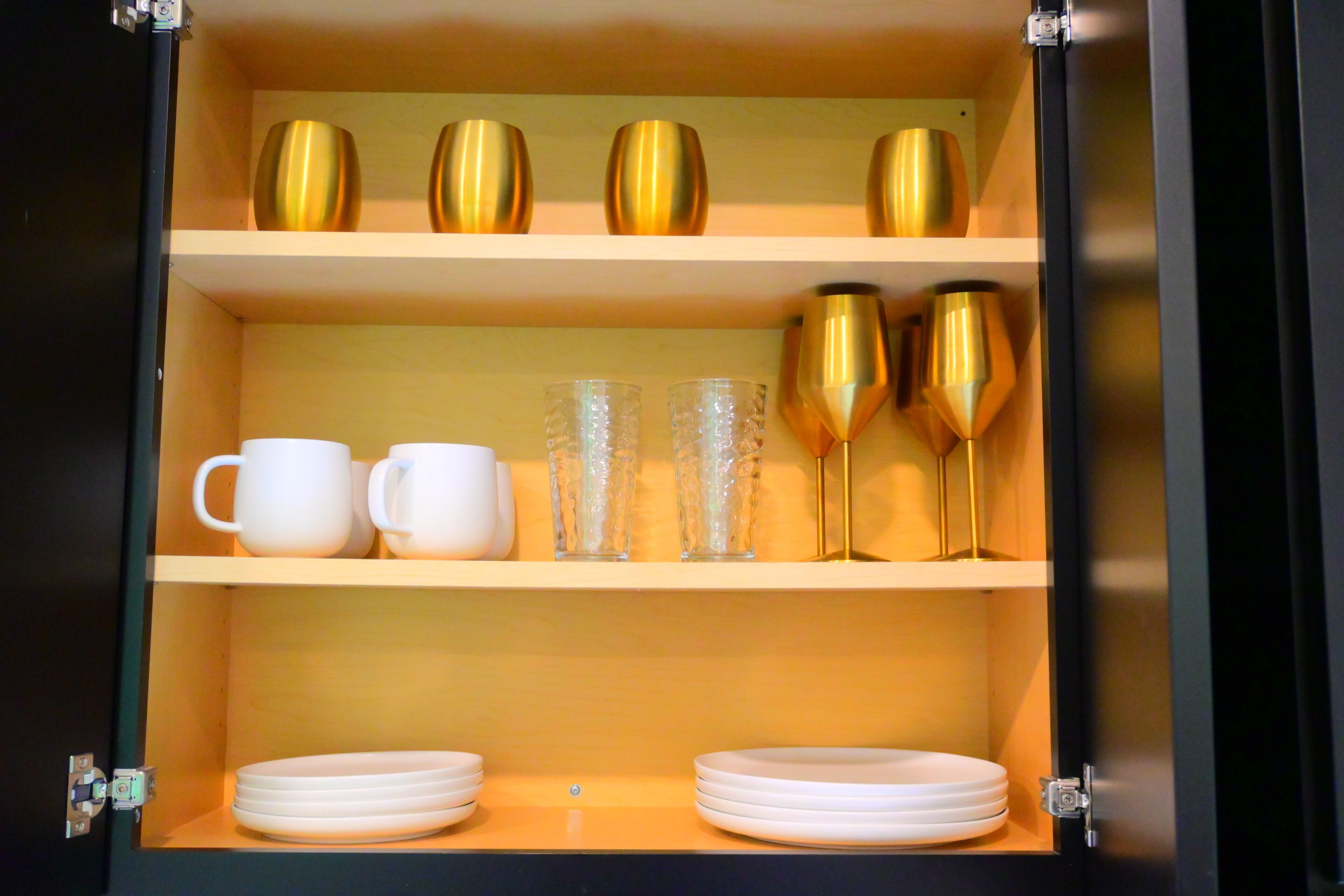 A cabinet with shelves containing white cups, clear glasses, and golden vases.