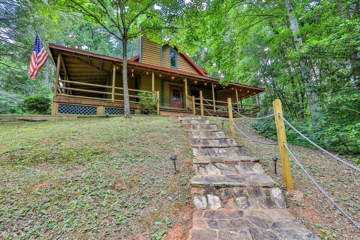 A rustic cabin with a stone staircase leading to a wooden porch, surrounded by lush greenery.