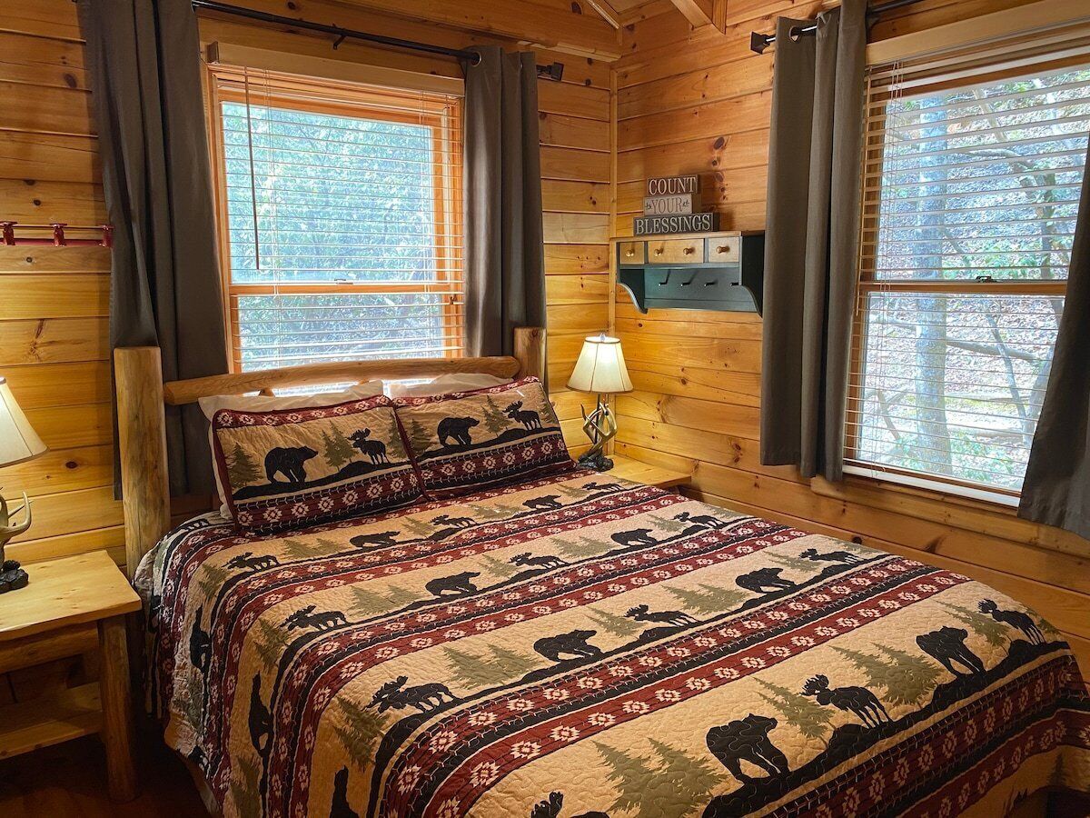 Un dormitorio de cabaña de madera con una cama adornada con colcha temática de osos, dos ventanas con persianas, una mesita de noche con lámpara y un estante montado en la pared con un letrero.