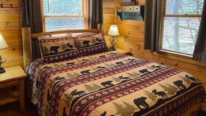 A wooden cabin bedroom with a bed featuring a bear-themed quilt, two windows with blinds, a bedside table with a lamp, and a wall-mounted shelf with a sign.